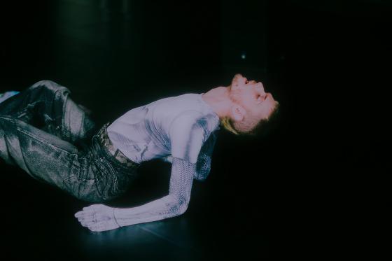 A person lies on their back on the stage floor, pushing the torso upward into a bridge and throwing the head far back. The fitted light top and dark trousers glow against the black space.