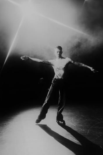 Black-and-white stage image: a single person stands with arms stretched out to the sides beneath a diagonal beam of light. The body casts a long shadow across the shiny floor.
