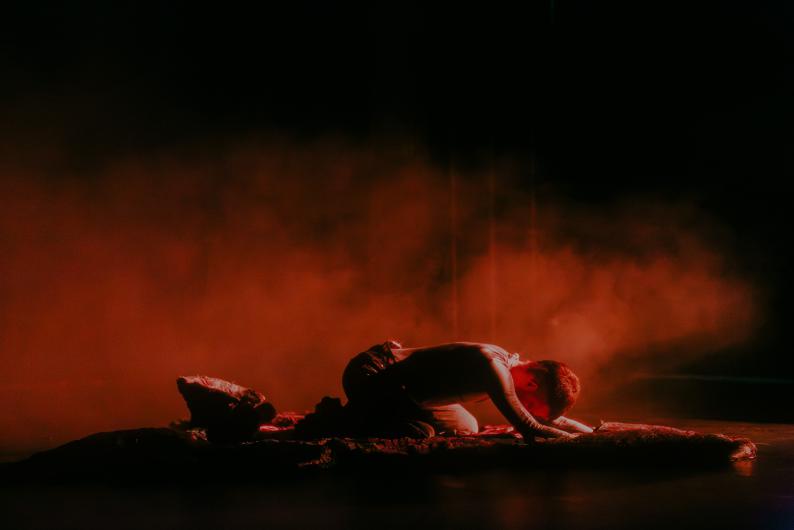 In red light, a person kneels collapsed over a low sculpture, forehead and arms close to the surface. Dense red haze drifts behind them through the black stage space.