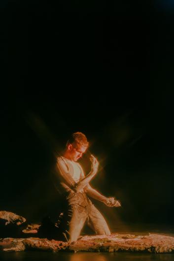 A person kneels on a flat surface and lifts one arm in front of the body as if examining or holding something. Warm light outlines the body against the black background.