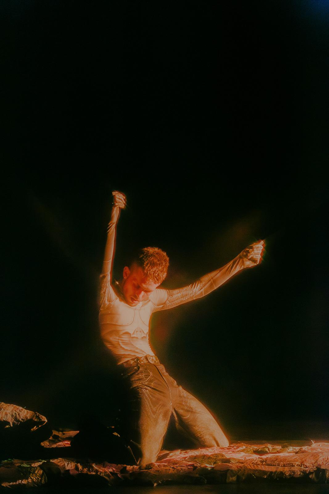 A person kneels with both arms raised on a low textured platform. Warm light isolates the figure while much of the stage remains black.