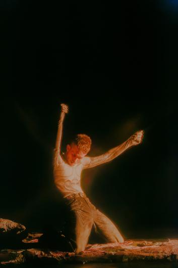 A person kneels with both arms raised on a low textured platform. Warm light isolates the figure while much of the stage remains black.