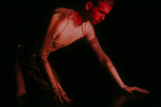 Close-up in red light: a person supports themself with both hands on the floor and looks downward. The light top and patterned sleeves stand out sharply from the black background.