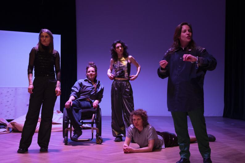 The five performers stand, sit, and lie together as a group. L speaks and gestures, I listens attentively, R and F gaze into the distance, T glances at R with a smirk. Five white bodies are connected, linked together. Behind them, screens and beanbags, drenched in pink-purple light.