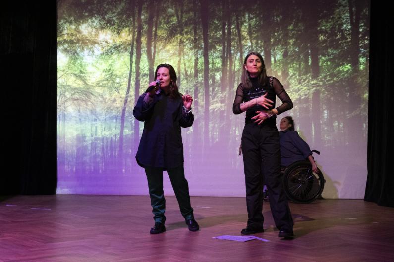 An enchanted forest. Mystical atmosphere, warm light. L speaks into a microphone, inviting the audience to a shared dance. R standing next to L, is signing. T in the background leaning back with pleasure.