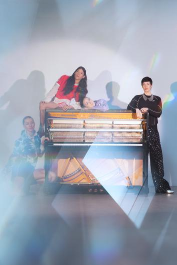 Four people pose around an upright piano with its inner mechanics exposed in an empty studio. One person in a red sports top and shorts sits on top of the piano, another person in a pastel lilac outfit lies relaxed across the lid. In front of the piano, a person in a blue-white tie-dyed hoodie and shorts kneels on the left, while a person in a black top and wide black trousers with white dots leans on the right side. Soft light and prismatic reflections cast a shimmering veil over the scene.