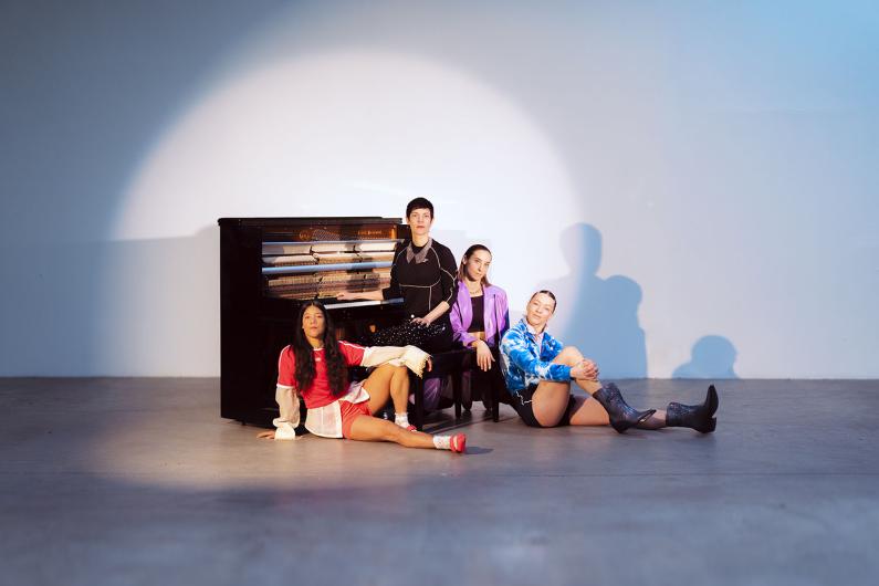The four people sit in a relaxed formation in front of a closed upright piano. The person in red sportswear sits on the floor to the left with legs stretched out. On the piano bench sits the person in a black top and dotted trousers, next to a person in a lilac blazer and dark trousers. On the right, a person in a blue hoodie and shorts sits with knees drawn up. A smooth spotlight surrounds the group and the piano against a plain studio wall.