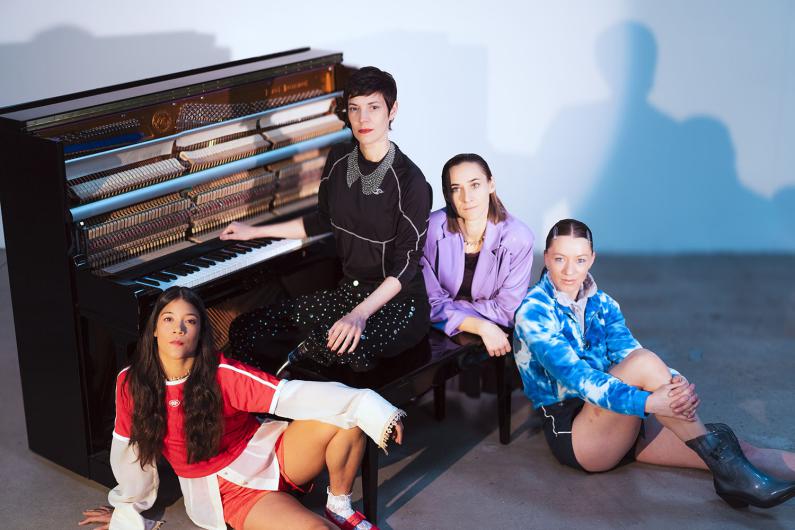 A closer view shows the four people closely grouped around the piano. In the front left, a person in a red sports top and shorts sits on the floor leaning against the instrument. On the piano bench, a person in a black top with a decorative collar sits beside a person in a lilac jacket and dark trousers. On the right, a person in a blue tie-dyed hoodie and shorts sits cross-legged wearing grey boots. The open piano with visible strings fills the background, while the light highlights faces and colourful outfits.