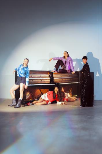 In a tall studio space, an upright piano stands near the bottom of the frame, surrounded by four people. On the left, a person in a blue tie-dyed hoodie, shorts and boots leans casually with one hand on the instrument. Sitting on top of the piano in the centre is a person in a lilac jacket and dark trousers with one knee raised. In front, a person in a red sports outfit lies almost fully stretched on the floor, while on the right a person in a black outfit with dotted trousers stands by the side of the piano. A wide circle of light falls from above, making the rest of the room appear almost empty.