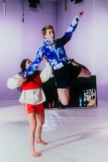 Eine Performer*in wird von einer anderen Person angehoben und balanciert mit ausgestreckten Armen in einem hellen Bühnenraum. Mikrofone hängen von der Decke.