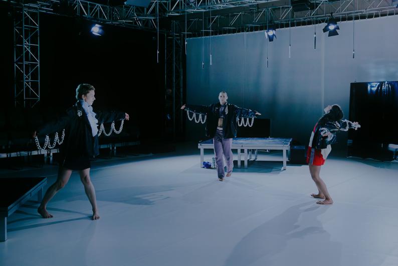 Drei Performer*innen stehen mit Abstand in einem großen Bühnenraum und bewegen die Arme seitlich. Sie tragen dunkle Jacken mit auffälligen, an den Ärmeln befestigten Ketten, die bei der Bewegung mitschwingen. Im Hintergrund sind eine Plattform und Lichttechnik sichtbar.