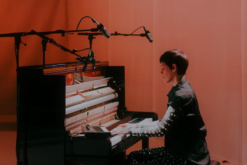 A person sits at an open piano and plays while several microphones are positioned above. Warm light casts the scene in orange tones.