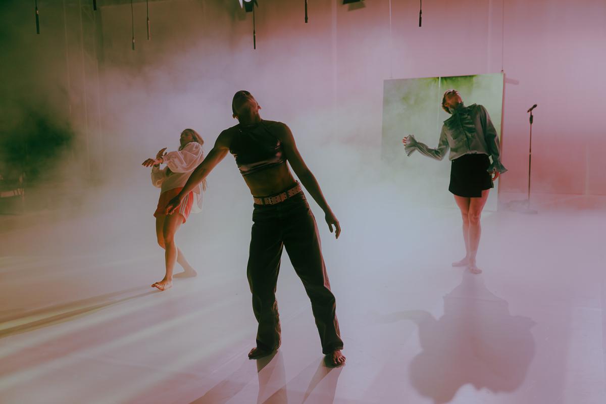 Three performers move through fog on a stage with colored lighting. Their bodies lean slightly backward as if drifting through the space.