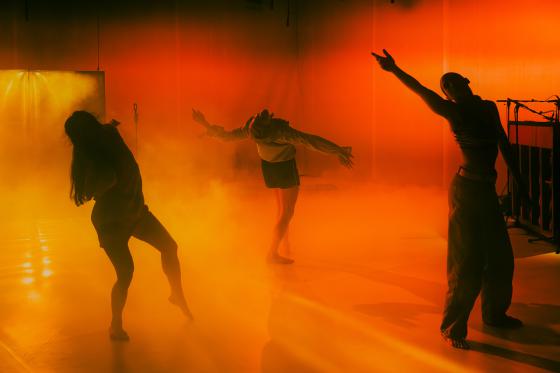 Three performers move through dense orange fog. Their arms are spread, and their bodies form expressive, open shapes.