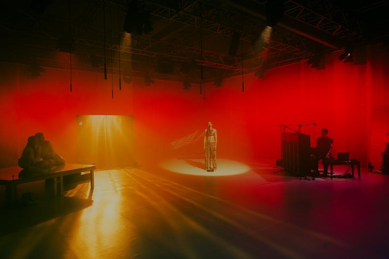 Eine weite Bühnenansicht zeigt mehrere Performer*innen, eine sitzende Person an einem Klavier und eine Person im Zentrum im Spotlicht. Rotes Licht und Nebel prägen die Atmosphäre.