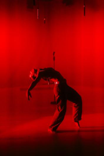 A performer moves alone on a stage fully immersed in red light. Their body bends backward, arms hanging loosely downward.