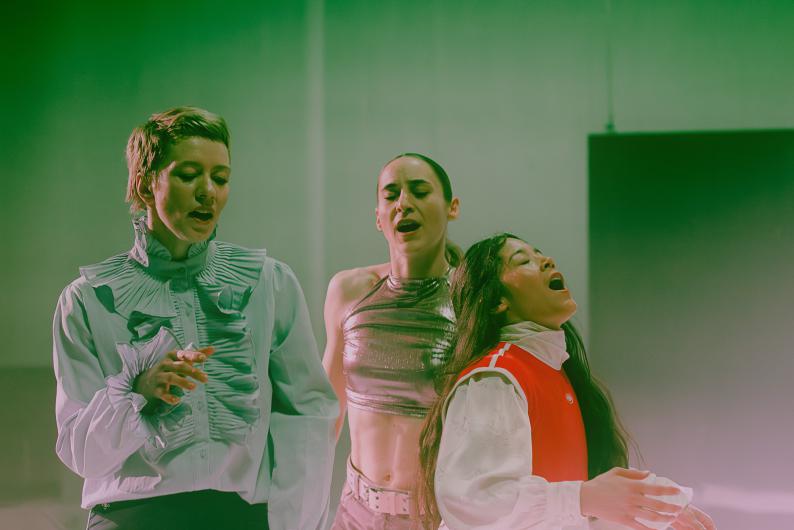 Three performers stand close together and appear to be singing or speaking simultaneously. Green light colors the scene.