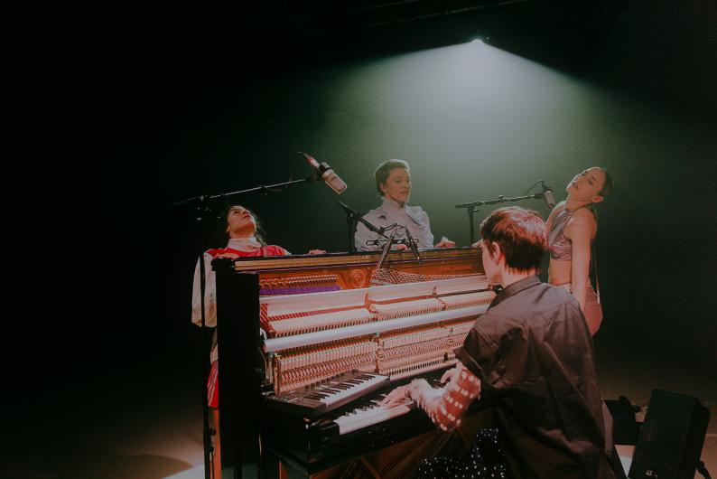 Three performers gather around an open piano while one person plays. Microphones hang above, and a spotlight illuminates the group.