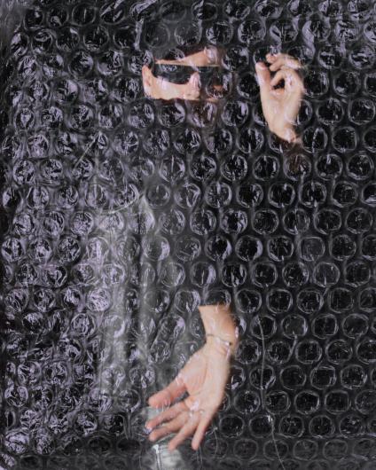 A person stands behind a continuous layer of transparent bubble wrap. The person is wearing a black cap and black sunglasses, which partially obscure the face. The bubble wrap covers the entire image and distorts the view of the body. Two of the person’s hands are visible pressing against the plastic: one hand reaches downward with slightly open fingers, while the other is raised and forms a delicate gesture with thumb and forefinger. The dark surroundings and the reflective, textured surface of the plastic create a sense of separation, protection, and simultaneous vulnerability.