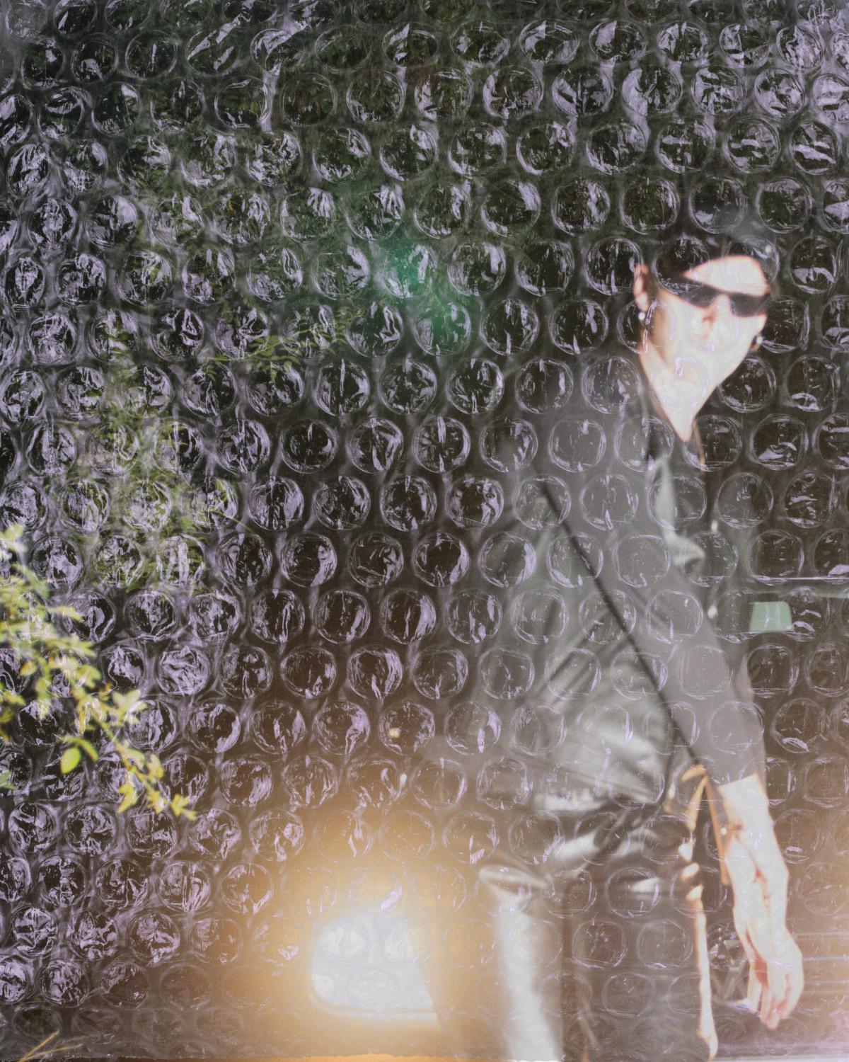A person stands behind a large sheet of transparent bubble wrap that covers the entire image. The person is wearing a black cap and black sunglasses; the face appears only faintly and is distorted by the textured plastic. The upper body is dressed in dark clothing. One hand hangs loosely downward and is visible through the bubble wrap. Blurred green plants and light reflections appear in the background and are mirrored on the shiny surface of the plastic. The interplay of transparency, reflection, and obstruction creates a sense of distance, shielding, and simultaneous presence.