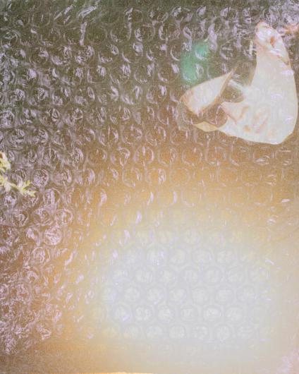 A person stands behind a large sheet of transparent bubble wrap that covers the entire image like a curtain. The round air pockets soften and slightly distort everything behind it, but the face is still visible: the person wears a black cap and black sunglasses. Light reflections and warm color gradients ranging from yellow to green shimmer through the textured surface, and blurred plant-like shapes are visible along the left edge of the image. The bubble wrap dominates the composition, creating a sense of shielding, blur, and distance.