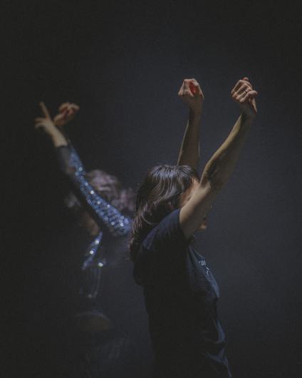 Two people stand one behind the other in a dark stage space with their arms raised. The person in front wears a dark T-shirt, while the person behind wears a sparkling long-sleeved top; both are caught by narrow beams of light.