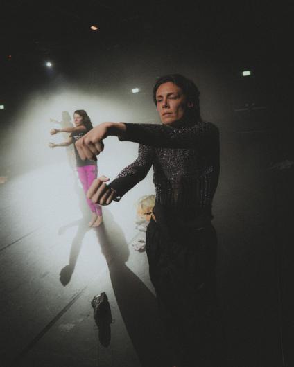 Zwei Personen tanzen in einem nebligen Bühnenraum. Im Vordergrund steht eine Person in einem glitzernden dunklen Oberteil mit angewinkelten Armen, weiter hinten bewegt sich eine zweite Person in pinker Hose im Gegenlicht.