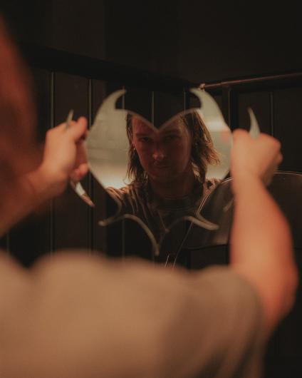 A person looks through two curved mirror shards or reflective objects held up with both hands. The reflection shows the face from the front, while the foreground and edges remain softly out of focus.