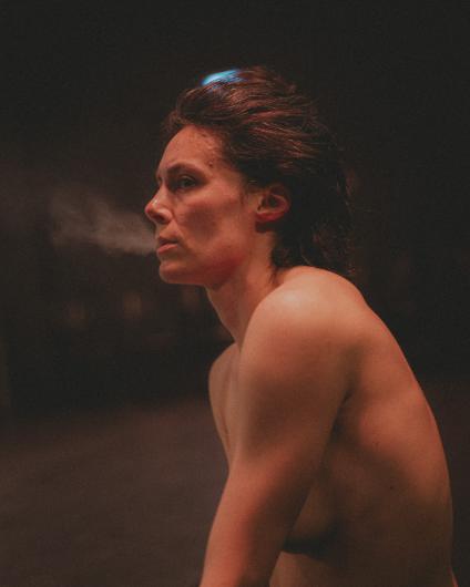 Close-up of a bare-chested person in warm, dim stage light, shown in profile facing left. A visible breath or mist drifts from their mouth. The background is dark and out of focus, with a small blue light glowing behind their head.