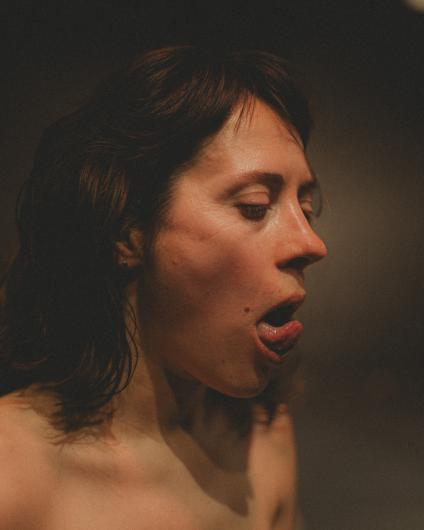 Close-up of a bare-chested person in warm light with their mouth slightly open and an intense facial expression. The background is dark and softly blurred, making the face and neck stand out sharply.