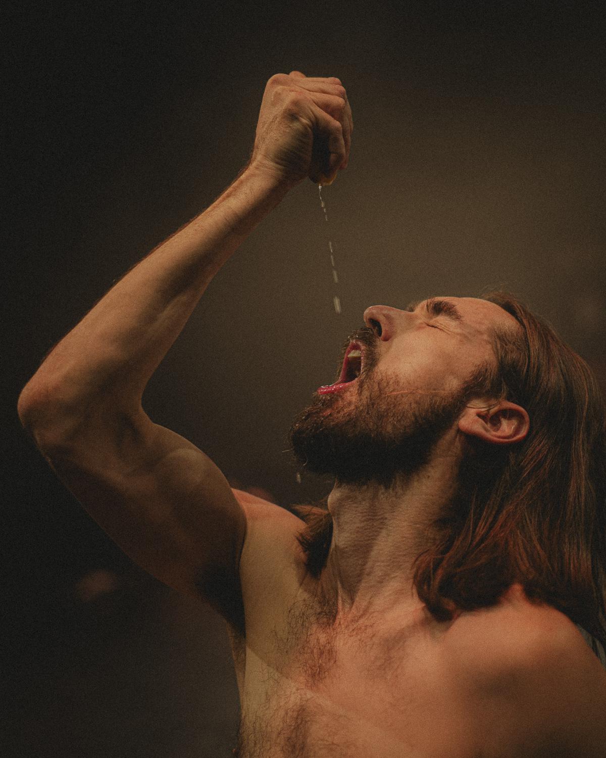 A bare-chested person with long hair and a beard tilts their head far back and lets liquid drip from a raised hand into their open mouth. Warm stage light highlights the arm, face, and torso against a dark background.