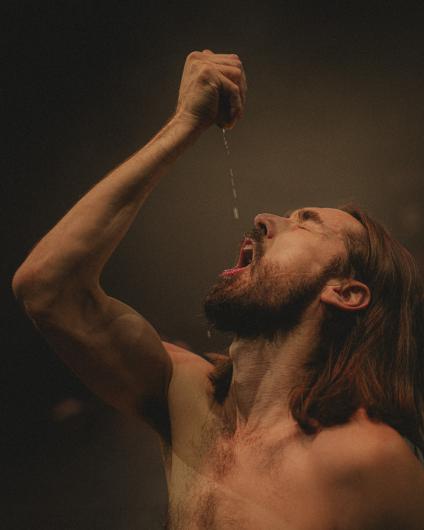 A bare-chested person with long hair and a beard tilts their head far back and lets liquid drip from a raised hand into their open mouth. Warm stage light highlights the arm, face, and torso against a dark background.