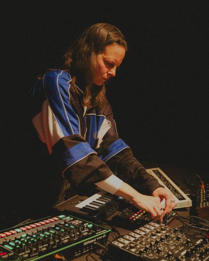 A person stands at a table of electronic music instruments and mixers, adjusting controls with both hands. They wear a sporty jacket in blue, black, and white, while the background remains dark.
