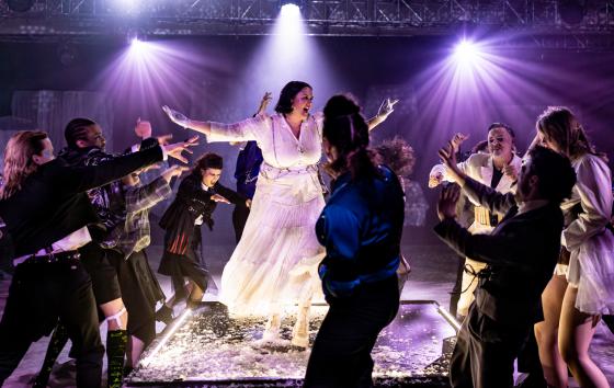 A person in a long white dress stands on a lit platform covered with white material at the centre of a surrounding group of performers. Around them, the other figures stretch their arms inwards, as if urging on, confronting or summoning the central figure. Purple stage light pours down from above, giving the scene a dramatic, almost ritual quality. Costumes range from dark suits to white, highly stylised stagewear. The image conveys intensity, motion and collective energy.