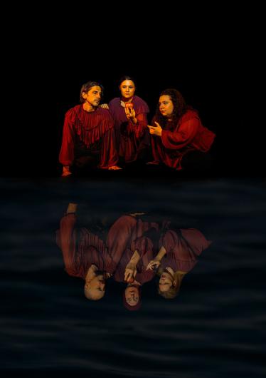 Three people in loose, dark red tops kneel close together against a black background. The person in the middle holds a red apple in one hand and looks forward, while the two people on the sides turn towards them – one of them points at the apple. Below the group, their image is reflected in a dark, slightly rippled water surface, where the three appear changed and partly in different poses.