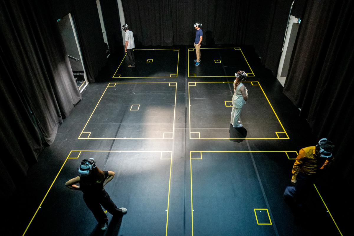 Several people wearing VR headsets stand in marked squares of a darkened room, separated by yellow lines.