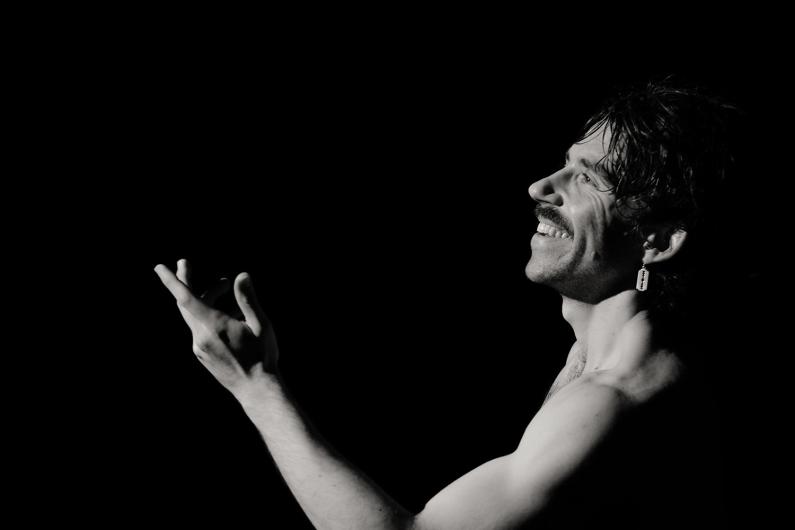 A person with a bare upper body, smiling in profile. The person raises one hand in an expressive gesture. The background is black.