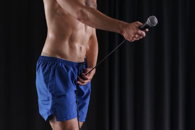 Nahaufnahme des Oberkörpers einer Person in blauer Shorts. Die Person hält ein Mikrofon in der Hand, der Hintergrund ist schwarz.