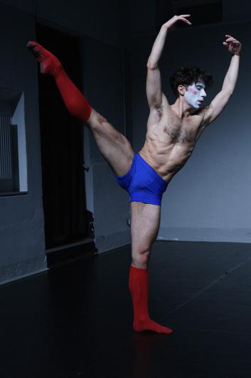 A person with a white-painted face and red lipstick stands on one leg with the other stretched high behind. They wear blue shorts and red knee-high socks, posing ballet-like on a dark stage.