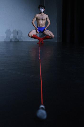A person with body paint squats deeply on a stage. A microphone with a red cable lies in front of them, the cable stretching to their hands. They look focused.