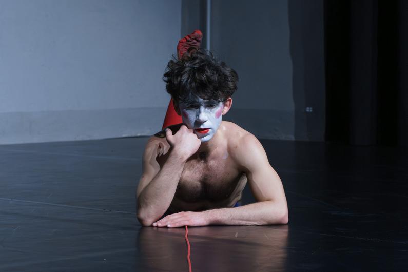 A person lies on their stomach on a stage, resting their head on one hand. Their face is painted white with red lips and cheeks, and a red microphone cable trails behind them.