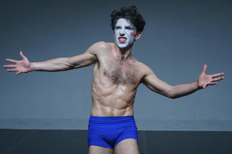 A person with face paint and a bare torso stands with arms stretched wide in an intense, almost theatrical pose.