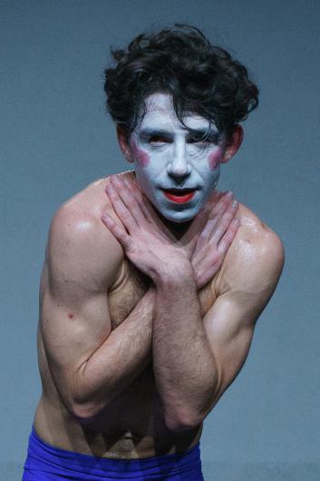 Close-up of a person with a white-painted face, red lipstick, and pink cheeks. Their arms are crossed over their chest, their expression vulnerable and direct.