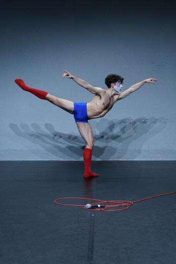 A person balances on one leg in a wide forward stretch. A red microphone cable loops on the floor in front of them. The light highlights the body’s tension.