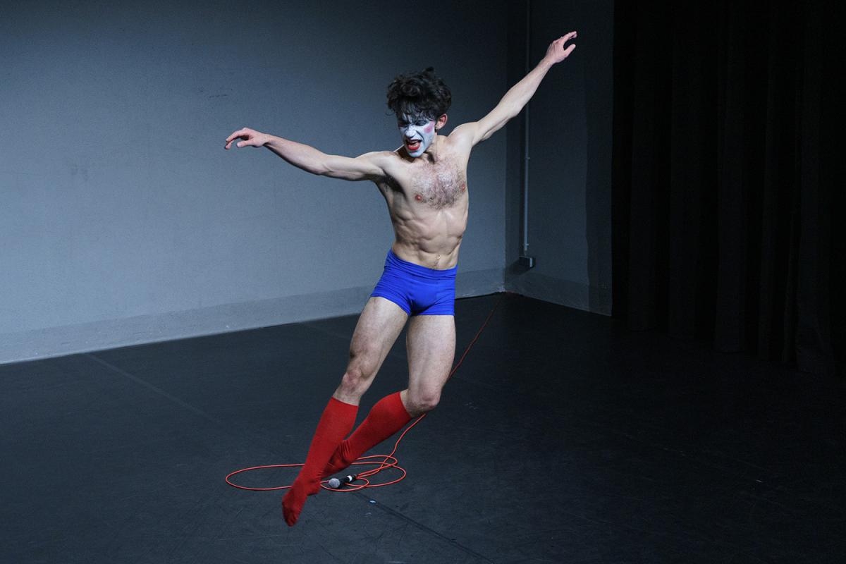 A person stands on a stage in an expressive dance pose with arms spread wide. They wear blue shorts and red knee-high socks, with a white-painted face and red lips. A microphone with a red cable lies on the floor. The background is plain gray.