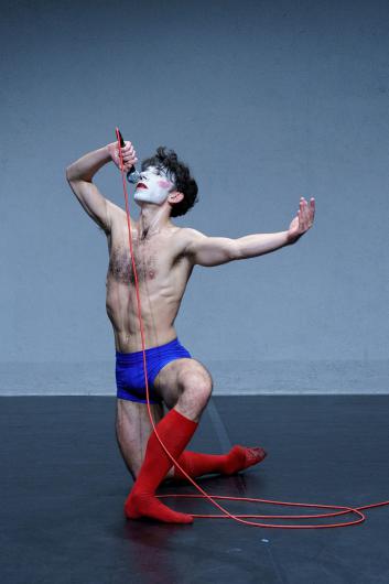 A person kneels holding a microphone, singing or speaking in an expressive pose. The red cable snakes across the floor.