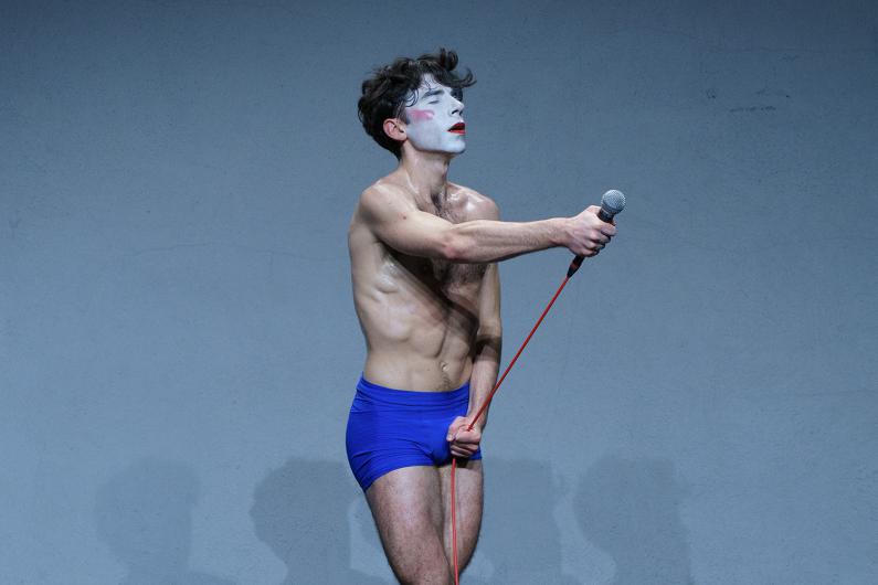 Eine Person steht leicht nach vorne gebeugt und hält ein Mikrofon mit rotem Kabel. Das Licht betont Schweiß und Bewegung. 