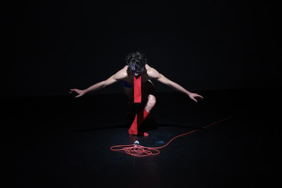 A person kneels in a pool of light while the stage around them remains dark. A microphone with a red cable lies in front of them, their body bent in a powerful pose.