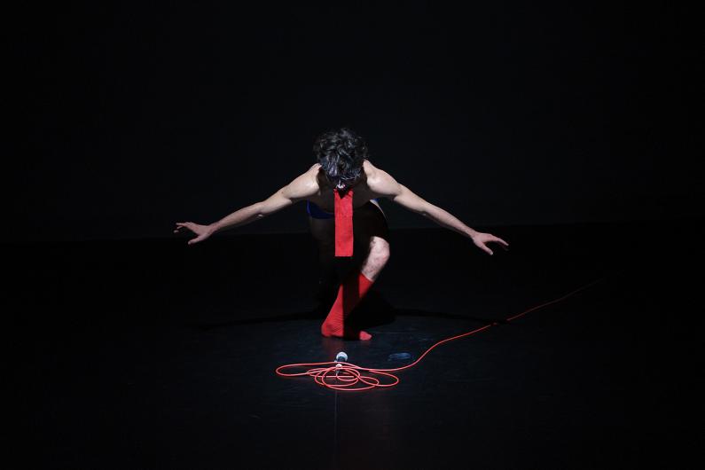 A person kneels in a pool of light while the stage around them remains dark. A microphone with a red cable lies in front of them, their body bent in a powerful pose.