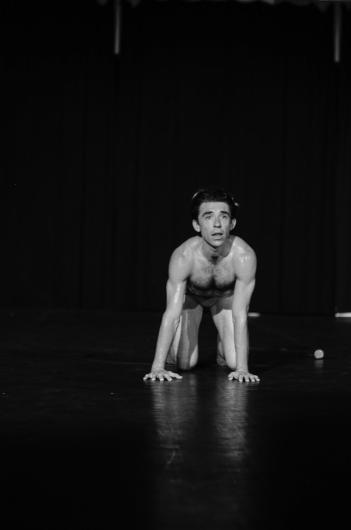 A person with a bare upper body kneels on all fours on a stage. The person looks forward, the background is dark.